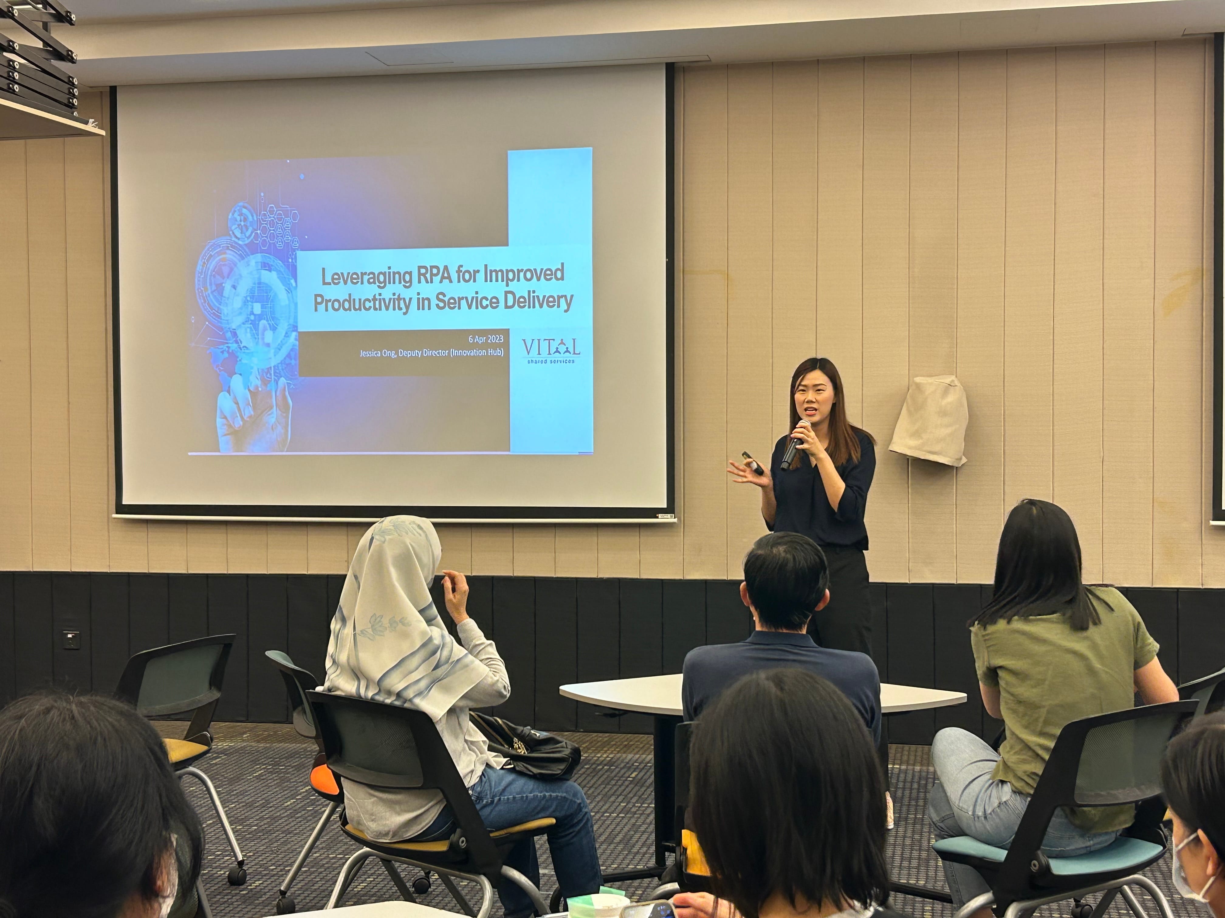 A woman speaks with a microphone before a projected slide titled Leveraging RPA for Improved Productivity in Service Delivery.